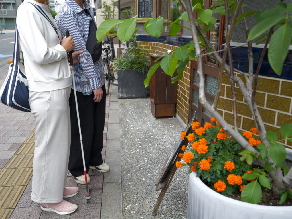 ガイドヘルパー木村さんが、利用者の白杖を持った女性を喫茶店の入り口に誘導しているところ。オレンジ色の花が入口わきのプランターに植えられている。