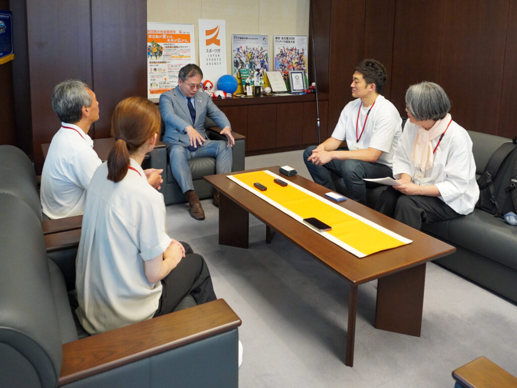 取材者4人に向け身振りを交えて話す河合さん。座った状態で両手を20センチ角程度に広げ、サッカーワールドカップを視察した時のデバイスのサイズを教えてくれている。
