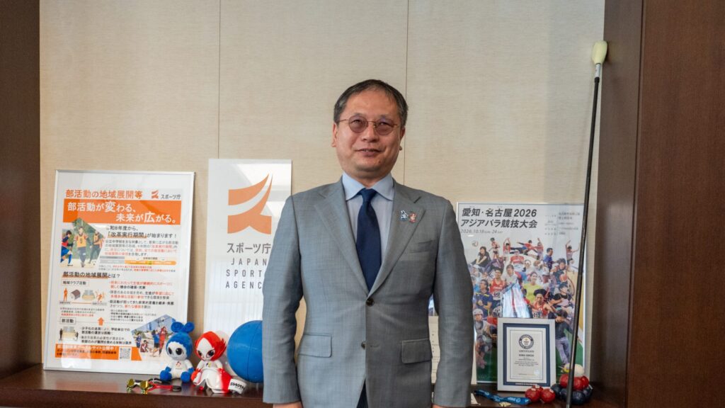 スポーツ庁内長官室の、ポスターなどが飾られた棚の前に、胸を張って立っている河合さん。
