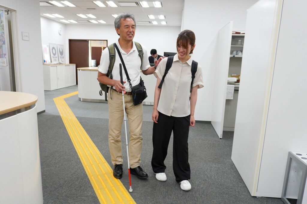 長官室に向かう室内の誘導ブロックの写真。パラアスリートの葭原さんが白杖を持ってガイドヘルパーと歩いている。
