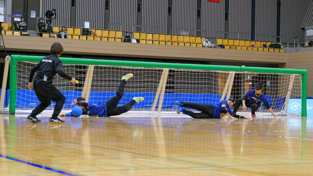 ゴールボール試合中の写真。3人の選手がゴール前で低い姿勢で飛び、ゴールを守っている様子。
