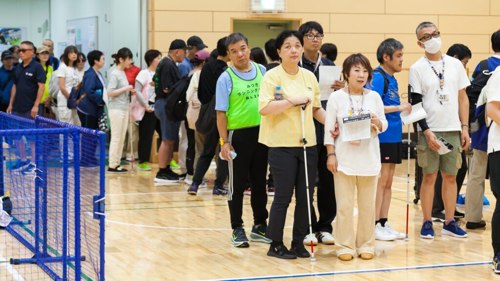 受付開始を待って並んでいる参加者の皆さん。全員動きやすい服装で、白杖を持った方、ガイドヘルパーなど100人近い方が並んでいる。