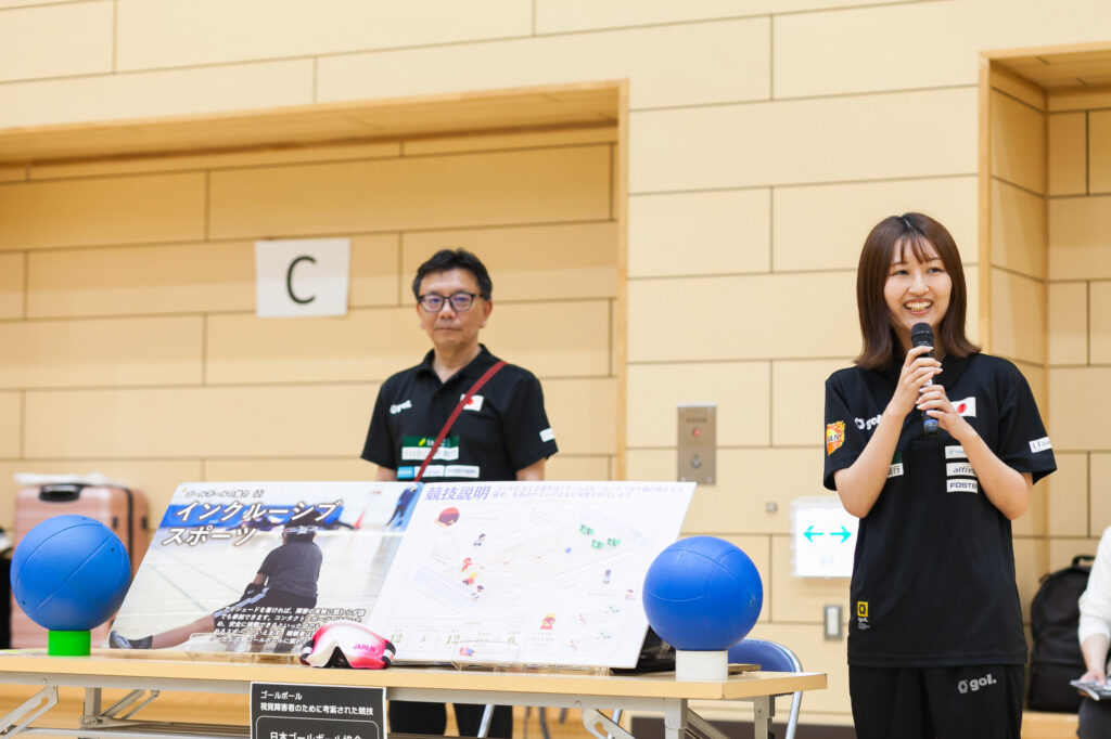 日本ゴールボール協会のスタッフが、マイクで笑顔で説明をしている。長机に説明パネルや実際のボールなどが設置されている。