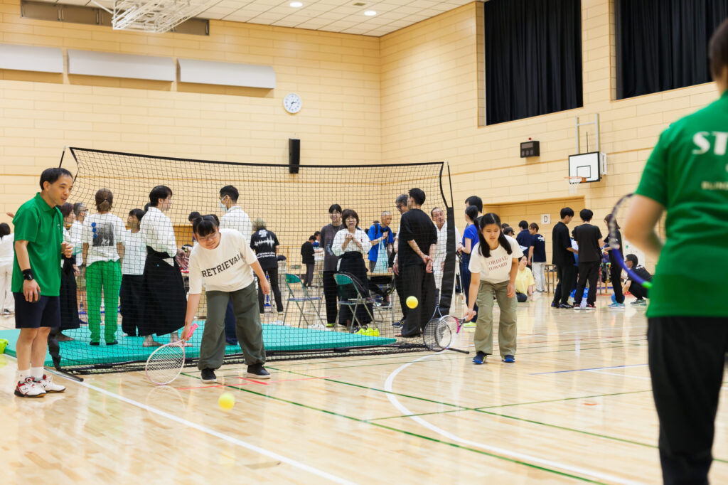 ラケットを右手に持ち、バウンドしているボールを打とうとしている体験者。説明スタッフが横に立ち見守っている。