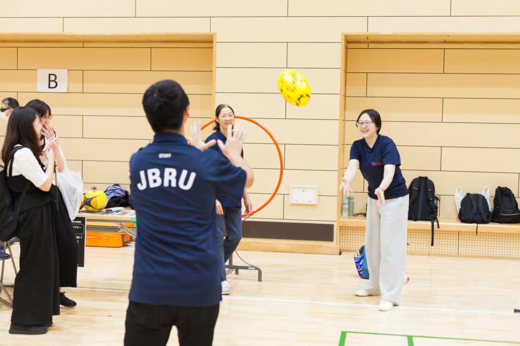 JBRUのロゴの入った紺色のポロシャツを着たスタッフが、体験者の投げたボールを受けようとしているところ。