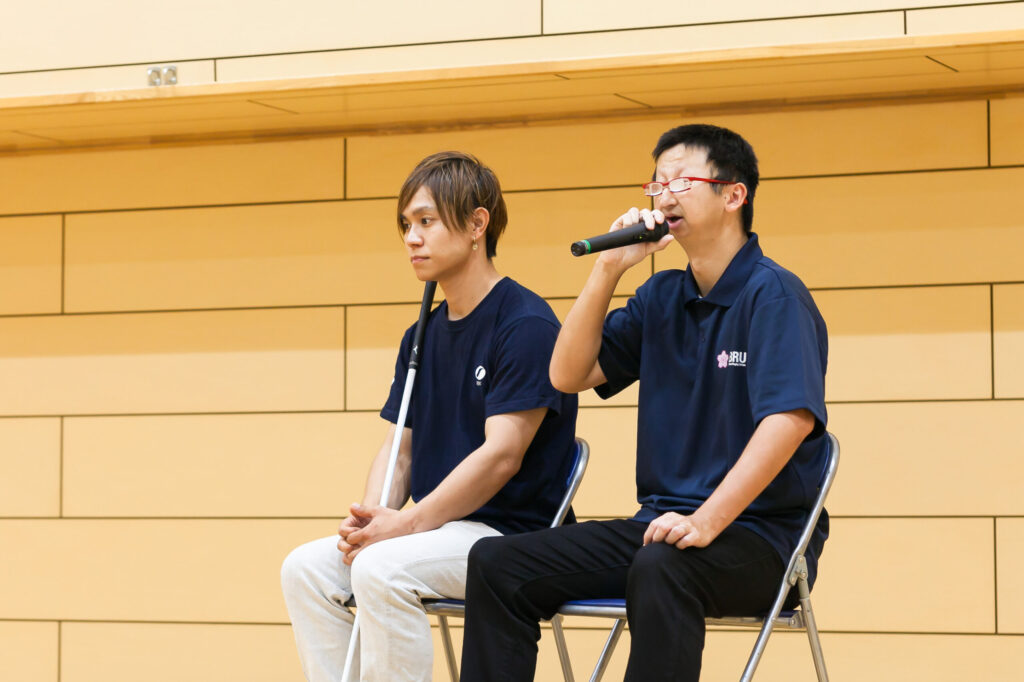 椅子に座り、右手にマイクを持って話している添田選手と、隣で手を膝の上に軽く合わせて聞いている濱ノ上選手。
