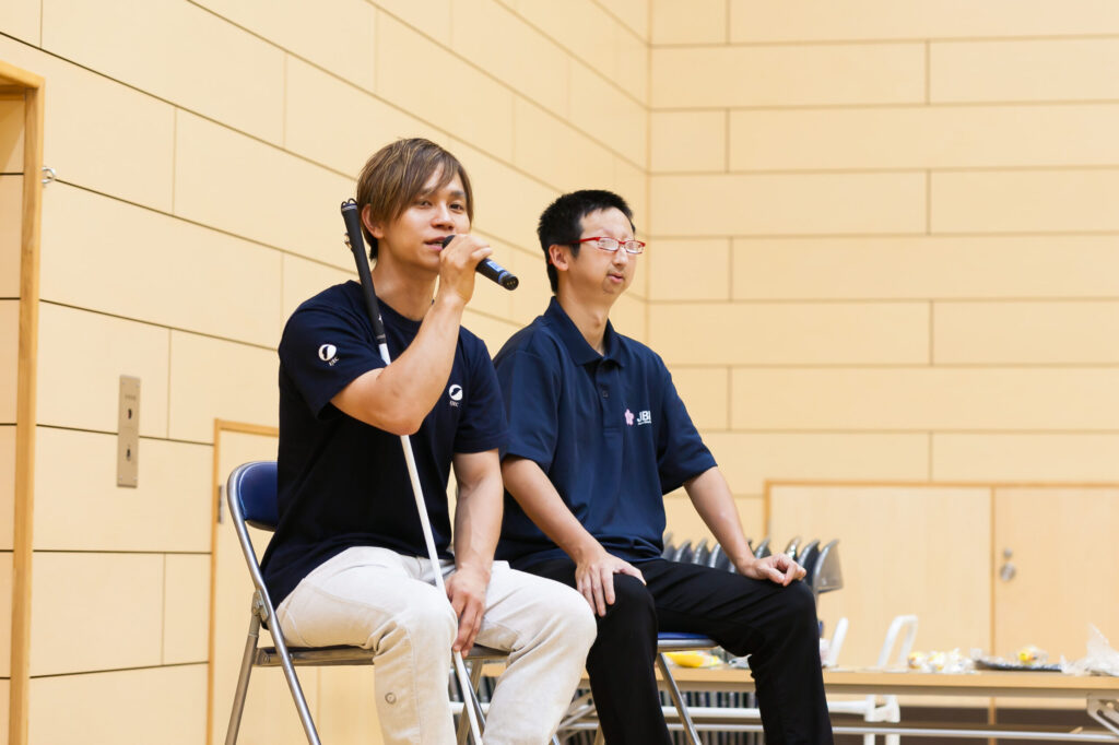 子に座って、右手にマイクを持ち話している濱ノ上選手と、ひざに手を多き聞いている添田選手。