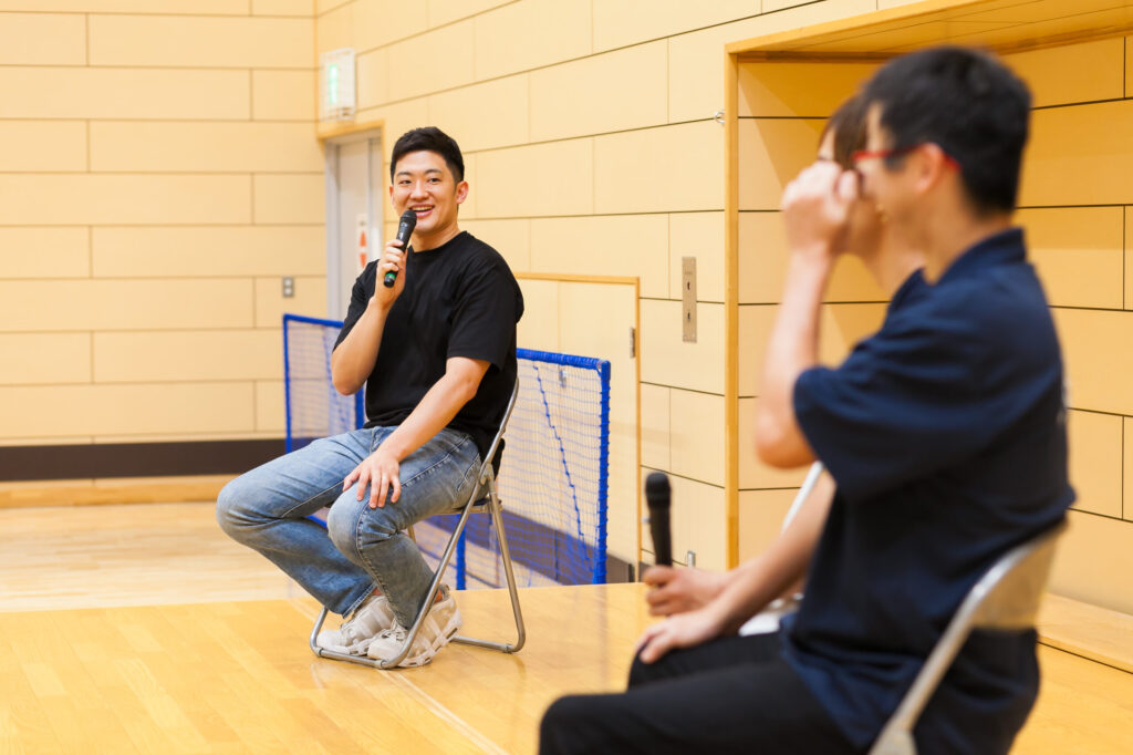 右手にマイクを持ち、濱ノ上選手と添田選手に向かって笑顔で質問している高橋。