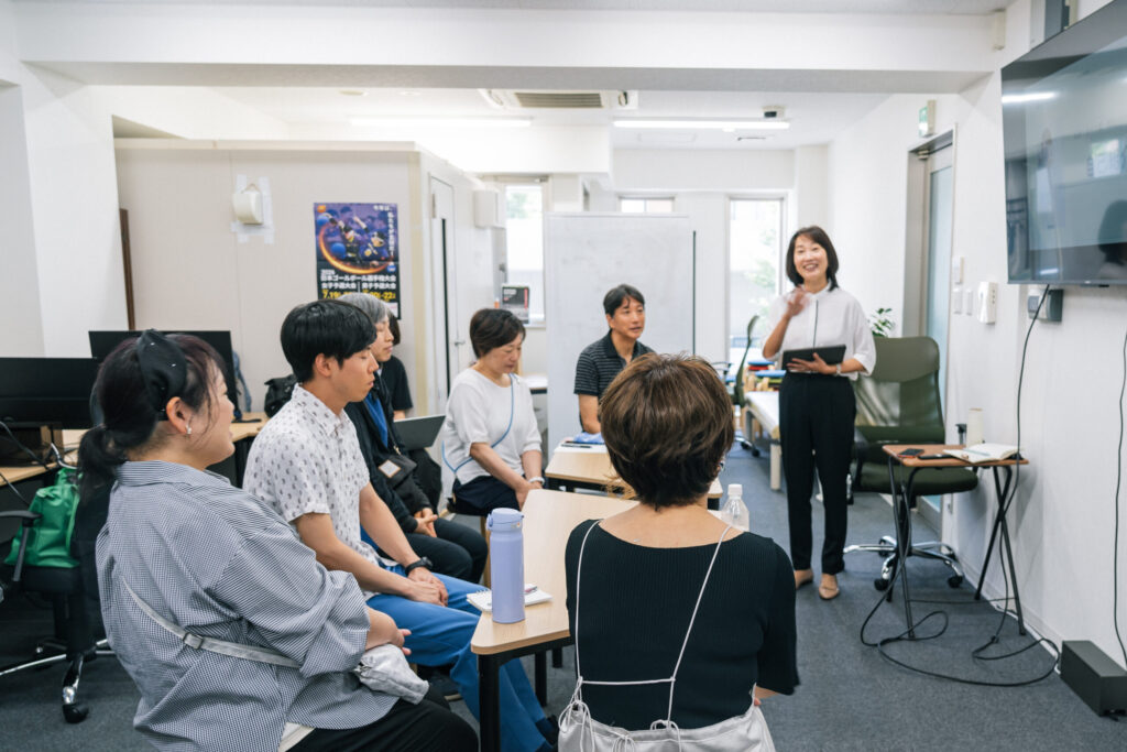 講師の内田さんが前に立ち、壁面の大型モニターを指さしながらにこやかに話している。6名の参加者が椅子に座って話を聞いている。